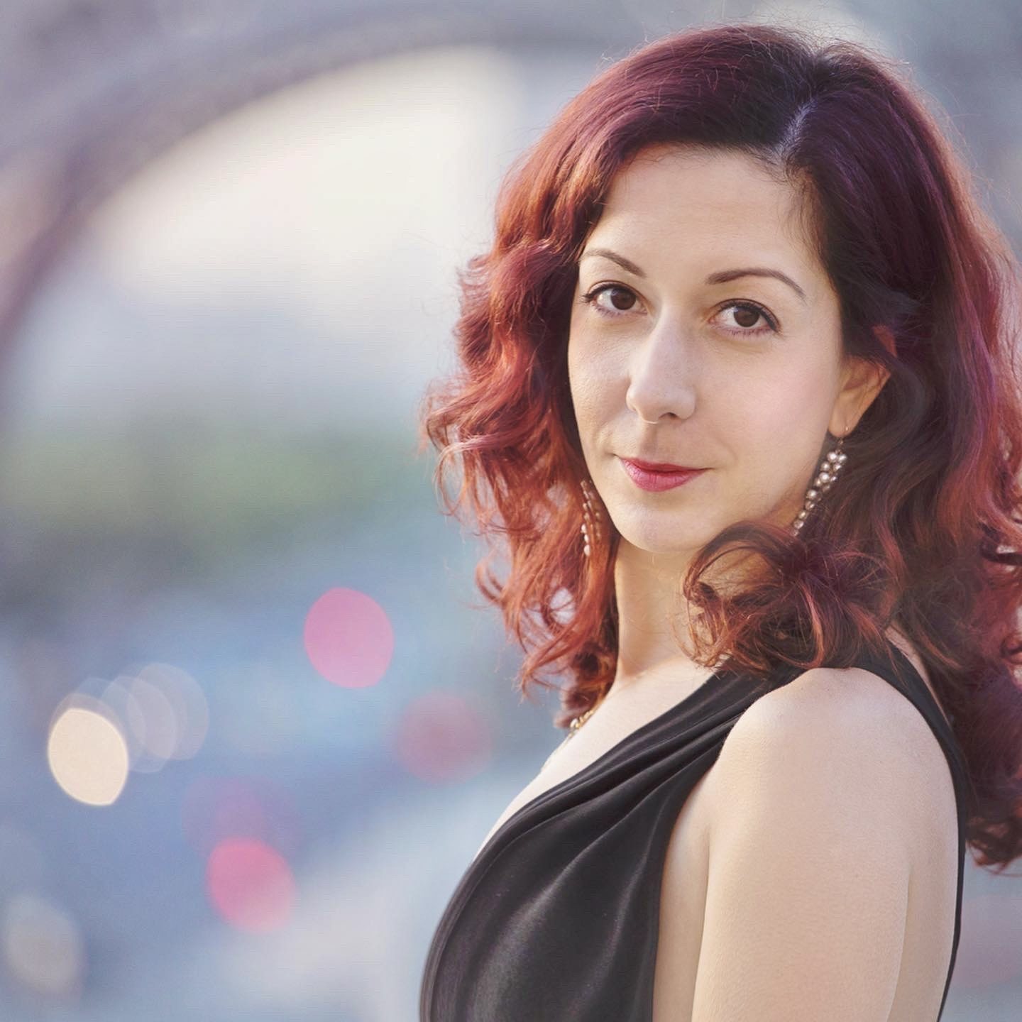 Close-up portrait of a red-haired woman in a black dress against soft city lights