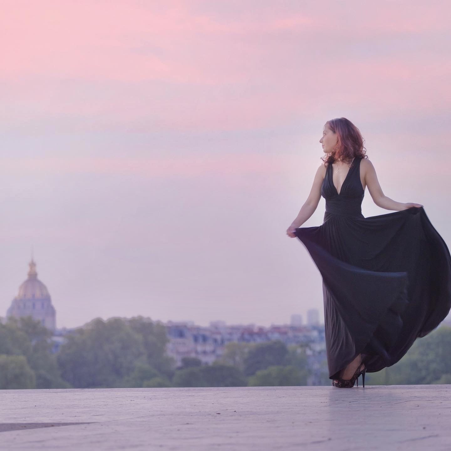 Woman holding her black gown against a soft pink sunset over the city
