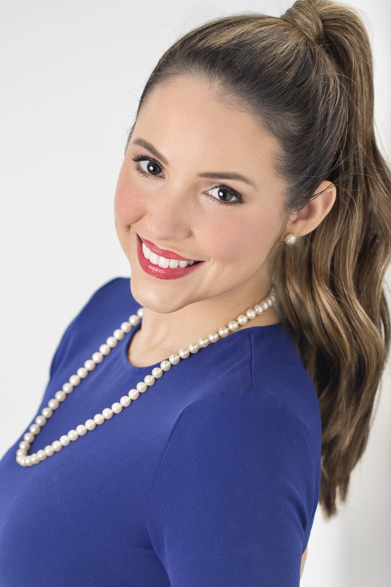 Smiling woman in a blue dress and pearls looking over her shoulder against a white background