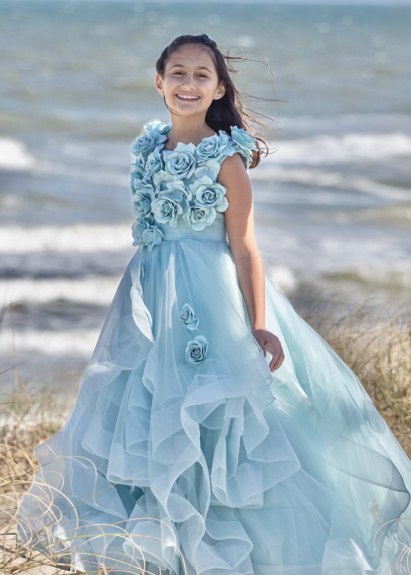 Smiling girl in a flowing pale blue gown beside beach grasses and ocean waves