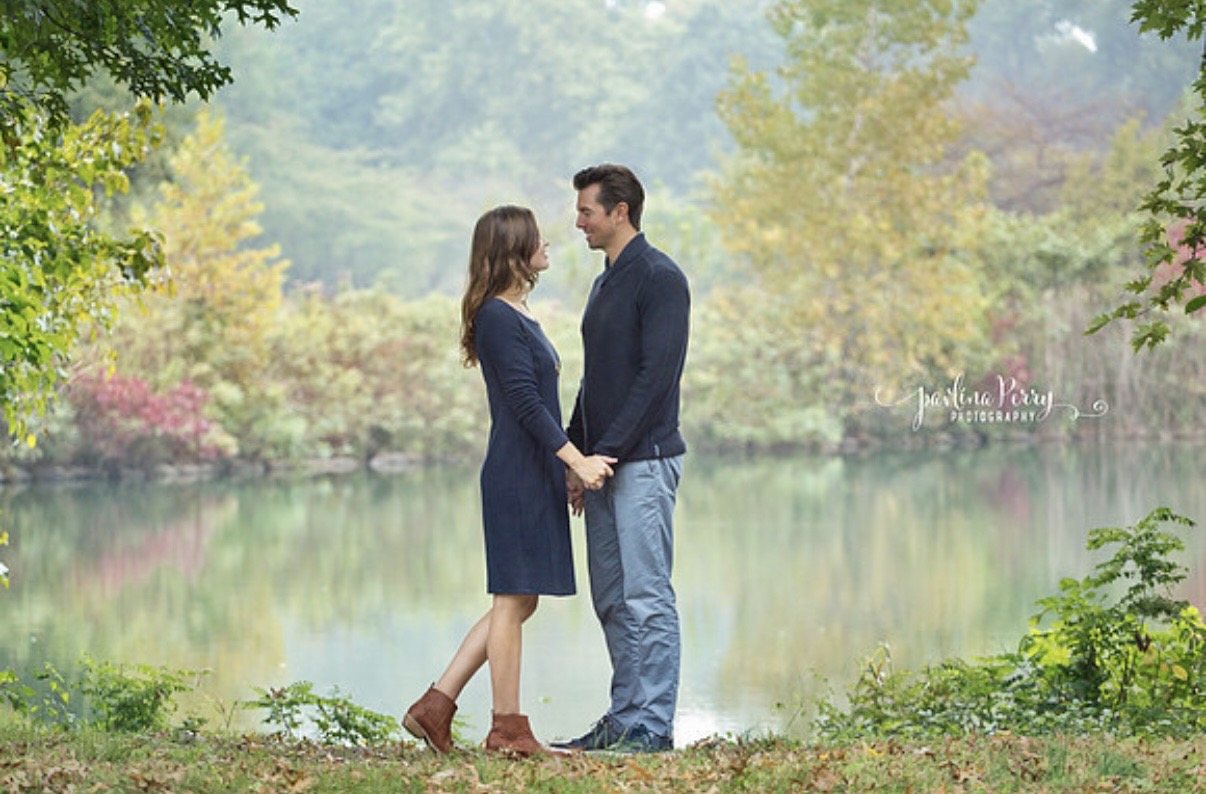 Couple holding hands beside a quiet lake surrounded by autumn foliage.