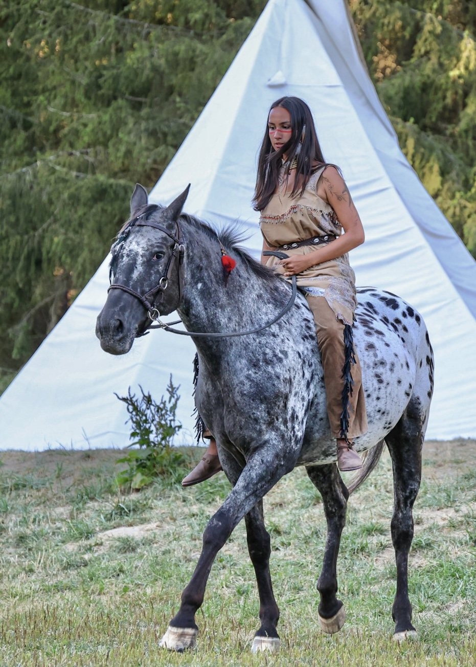Woman riding a spotted gray horse past a white tent at the edge of woodland