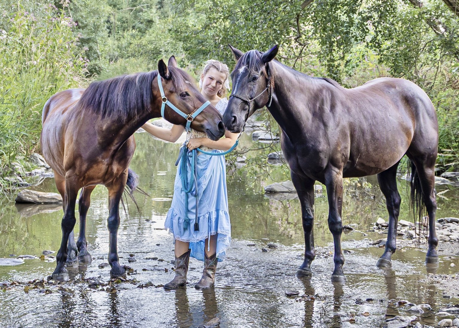 Woman in a blue dress and riding boots standing with two horses in a shallow creek surrounded by trees
