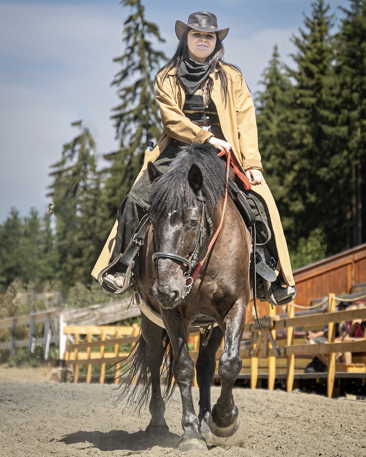 Western rider in a tan coat and black hat riding a dark horse toward the camera in a sunlit arena