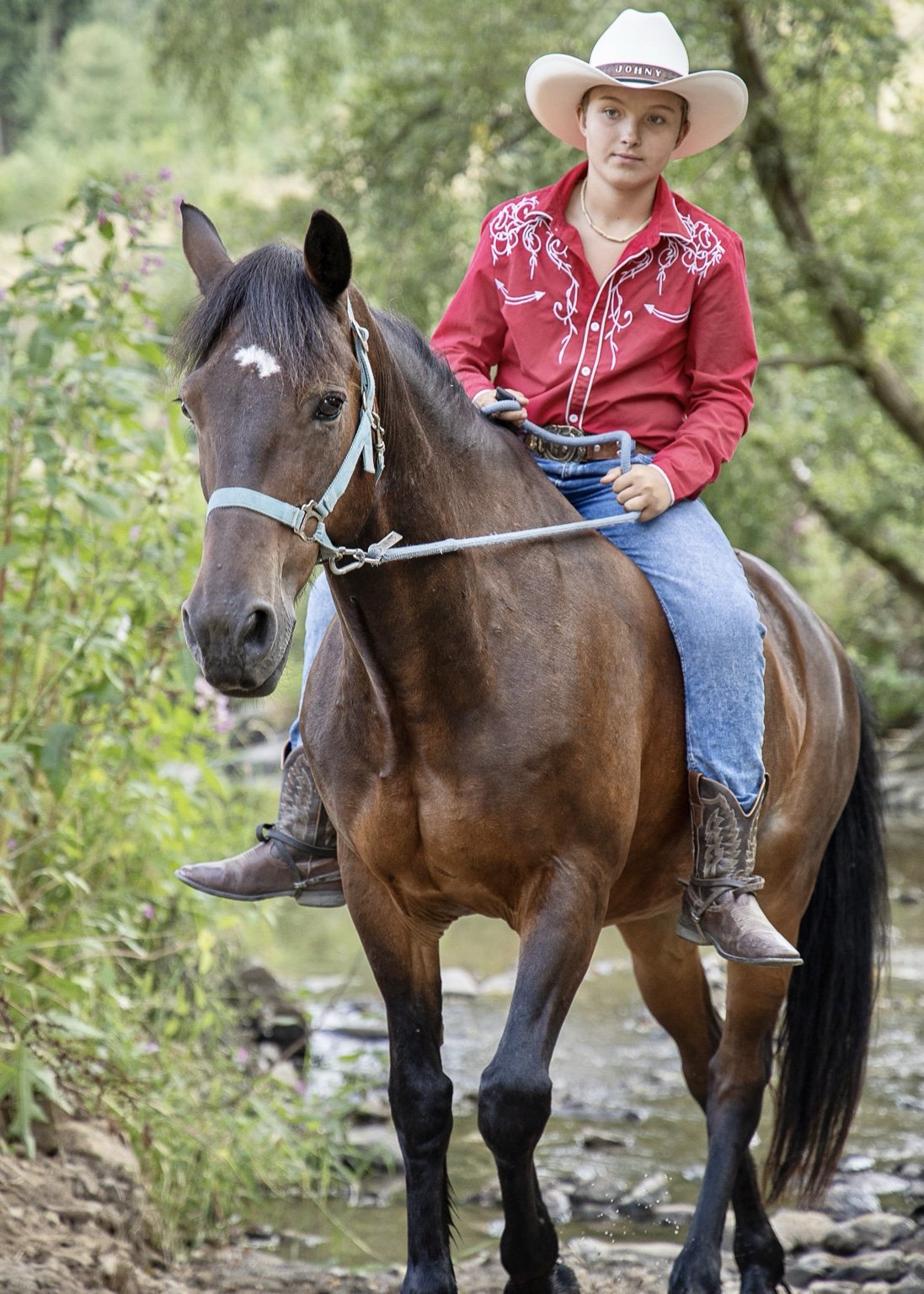 Boy in a red western shirt and white cowboy hat riding a brown horse beside a woodland stream