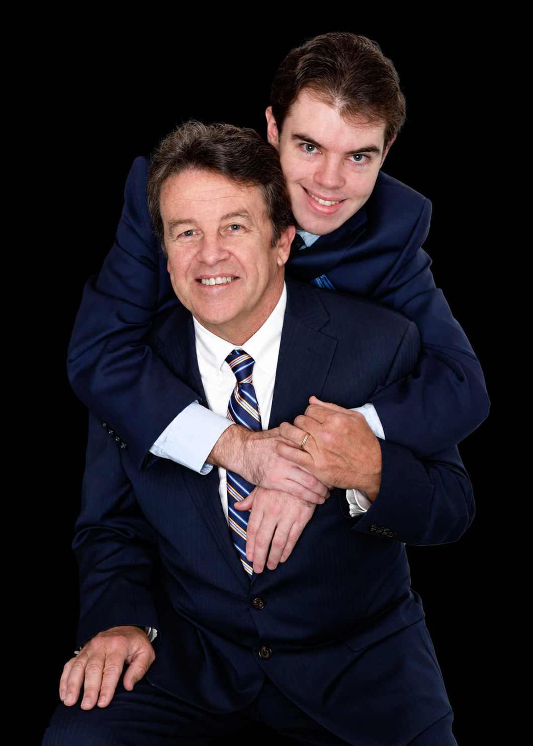 Grown son in a navy suit hugging his smiling father from behind against a black background