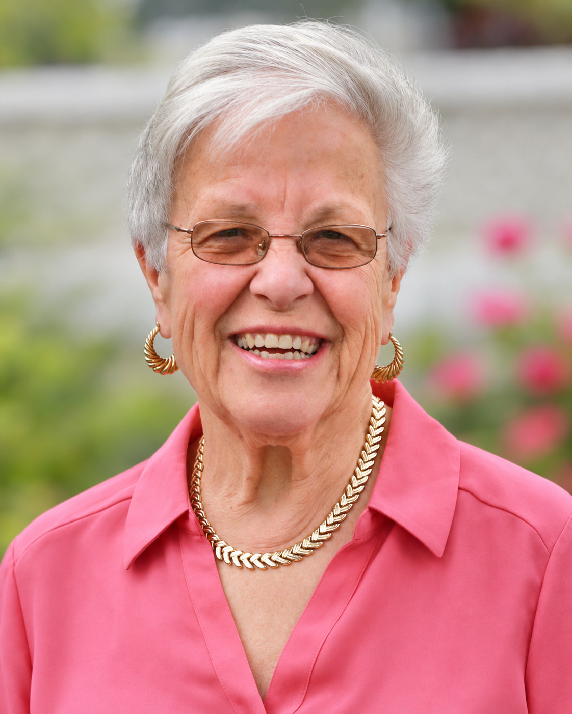 Smiling silver-haired woman in a pink blouse and pearl necklace with a garden behind her