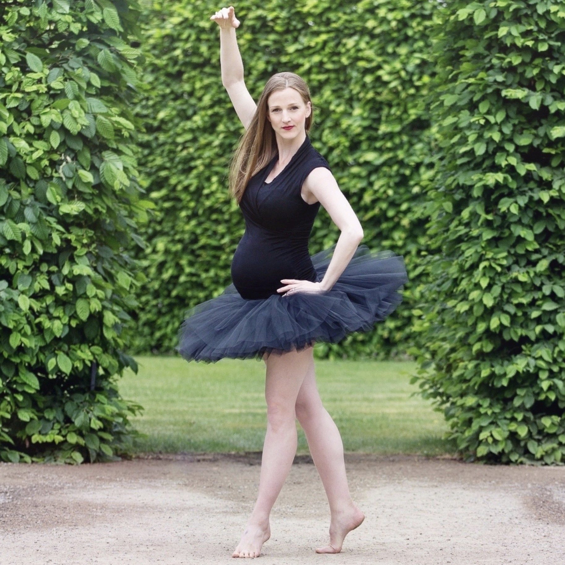 Full-length maternity portrait of a barefoot dancer in a dark tulle skirt, posing between leafy green hedges
