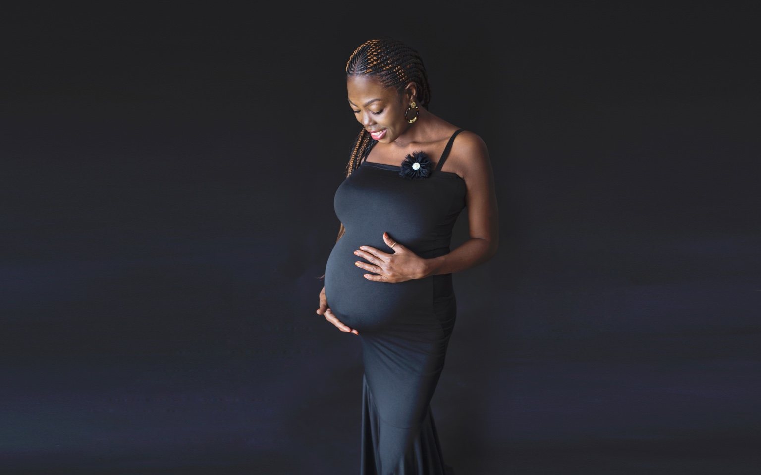 Maternity portrait of a woman in a dark gown looking down while gently holding her belly against a dark background
