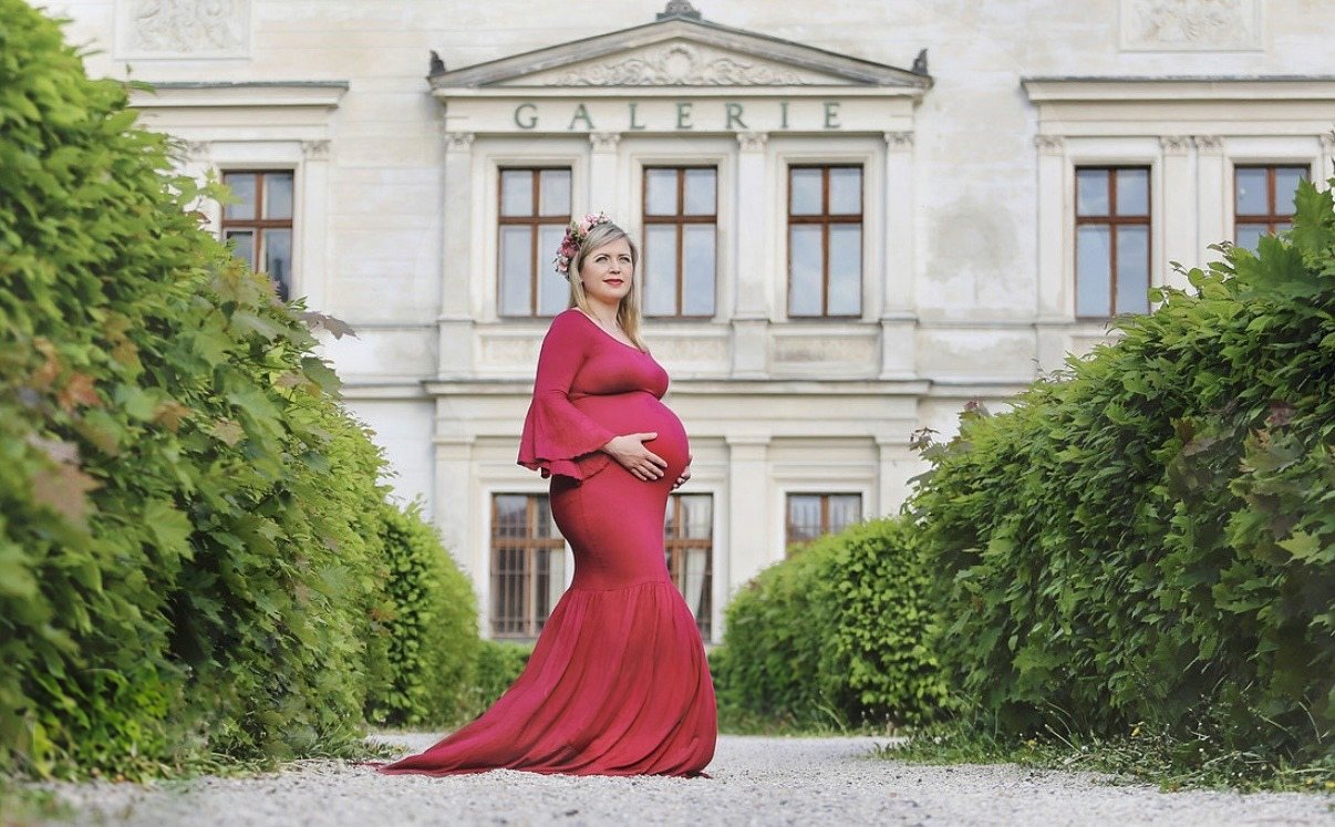 Maternity portrait in a flowing magenta gown between green hedges in front of a historic gallery.