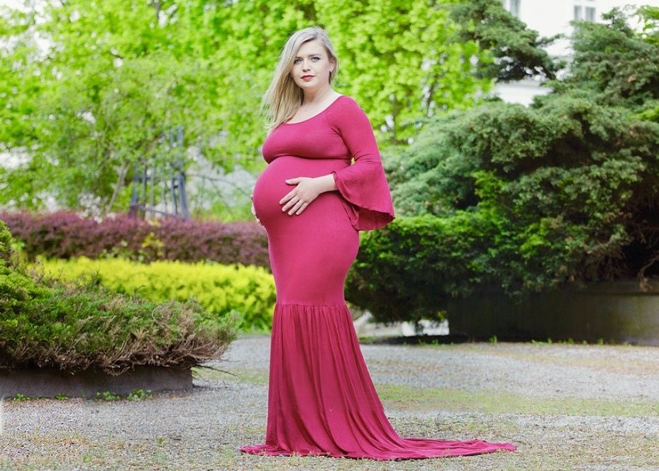 Maternity portrait in a full-length magenta gown in a lush green garden.
