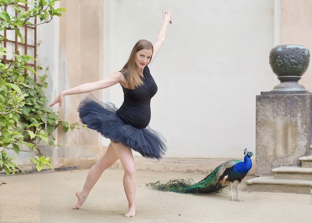 Maternity portrait in a dark tulle dress, dancing beside a peacock in a historic courtyard.