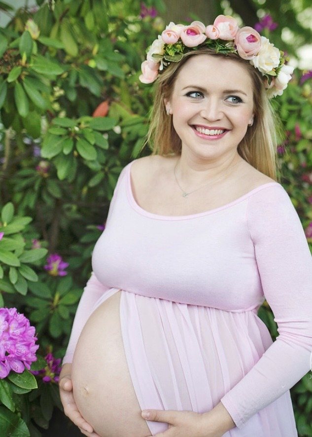 Smiling maternity portrait in a pale pink gown and floral crown among flowering shrubs.