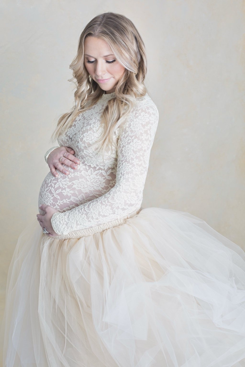 Expectant mother looking down and gently cradling her belly in an ivory lace top and flowing tulle skirt