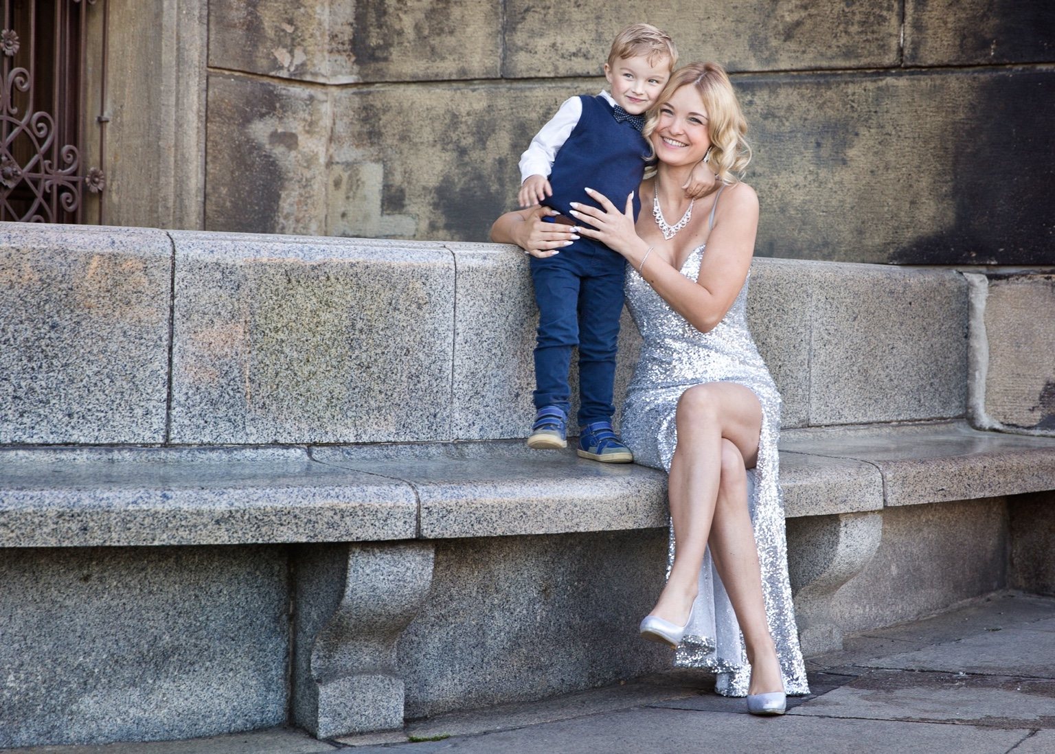 Mother in a silver gown seated beside her young son in navy against historic stone architecture.