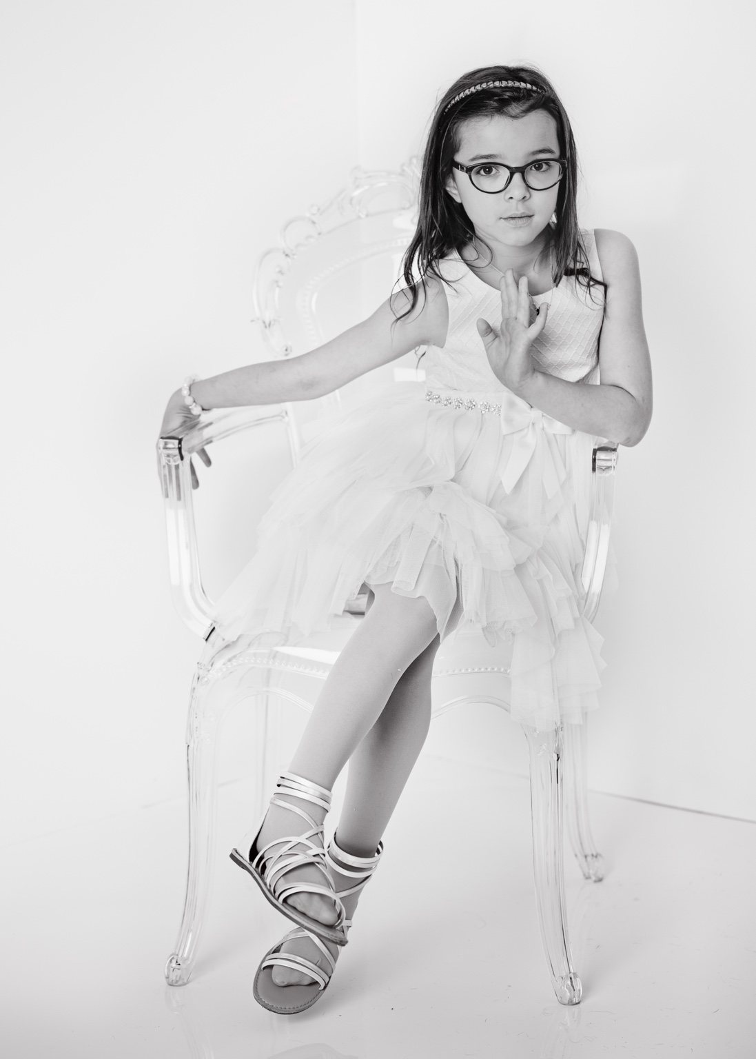 Black-and-white studio portrait of a girl with glasses in a white dress, seated on a clear chair