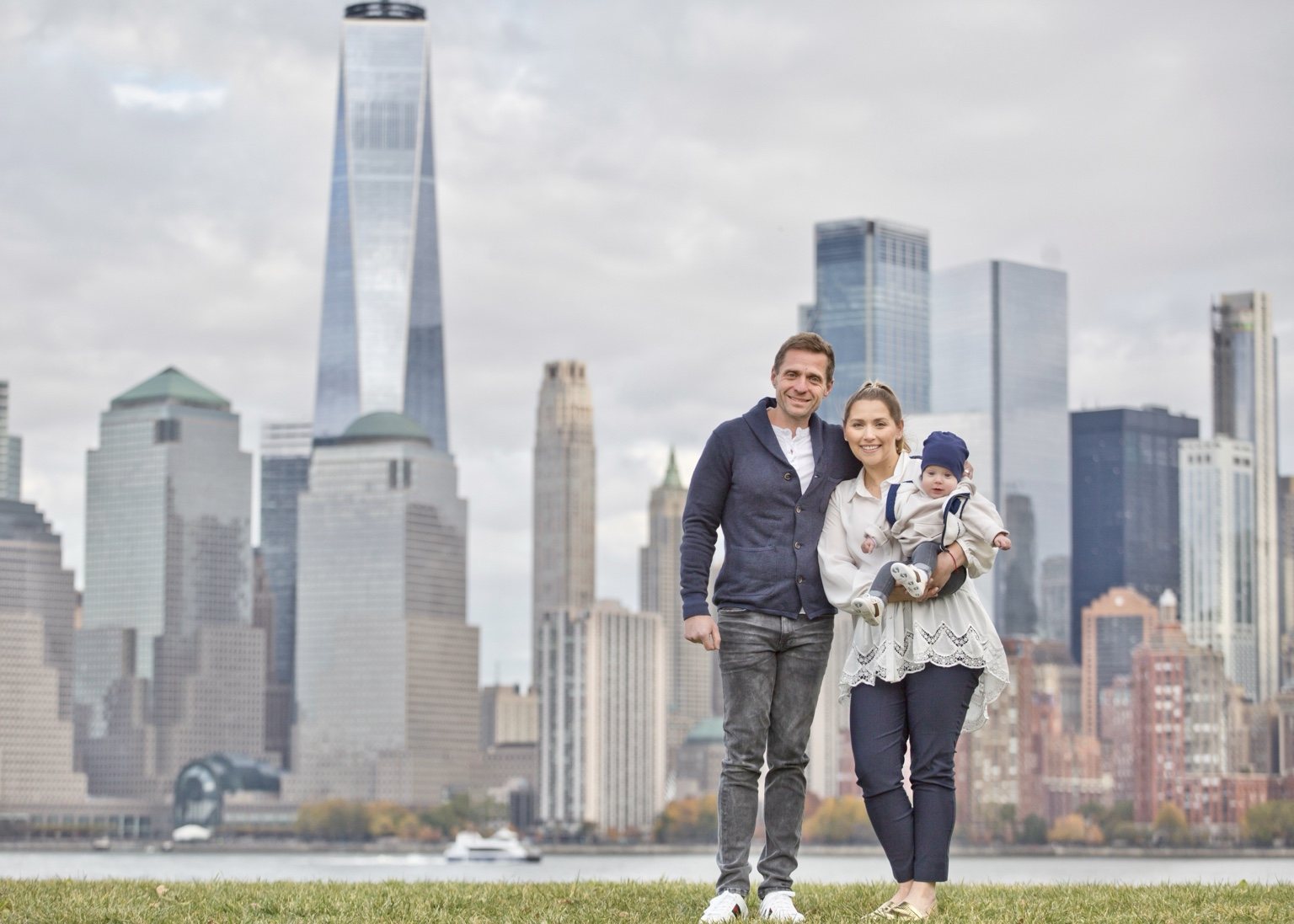 Parents holding their baby on the waterfront with the Manhattan skyline behind them.
