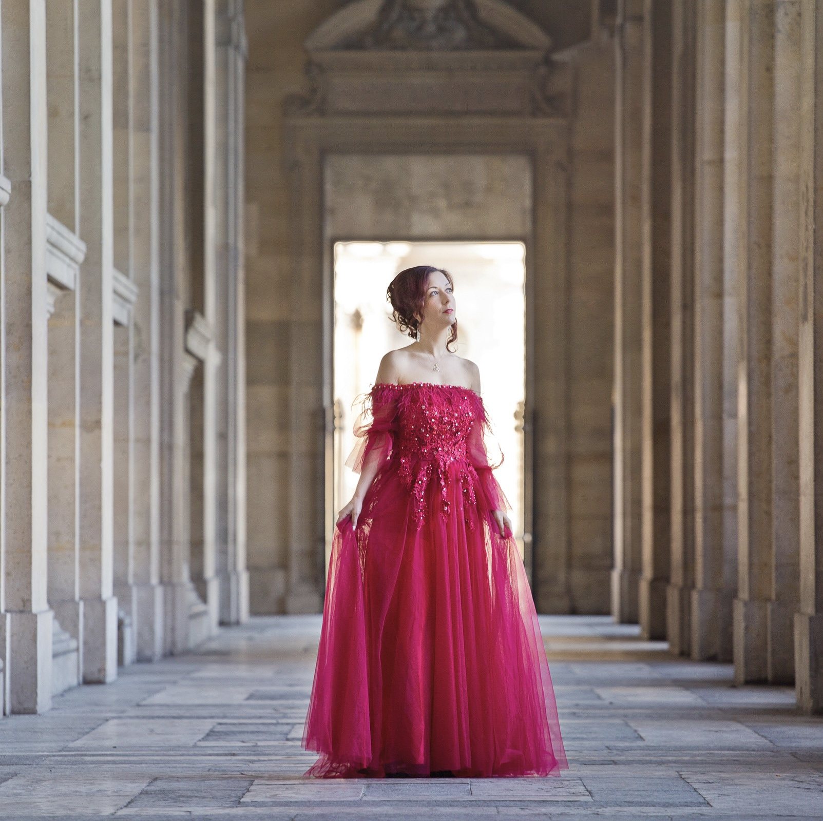 Woman in a vivid red gown standing within a stone colonnade