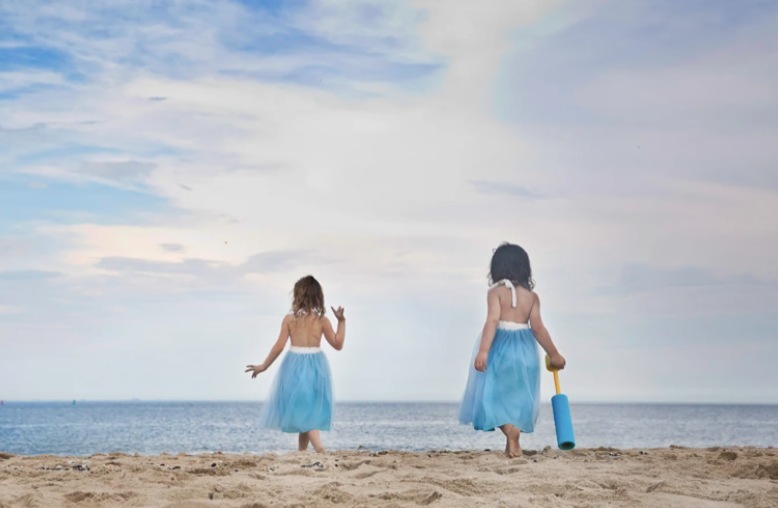 Two little girls in pale blue tulle skirts walking across the sand toward the sea
