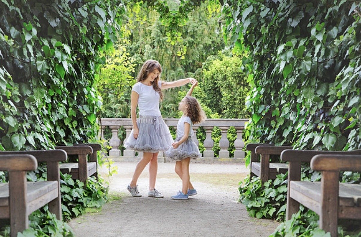 Two sisters in matching gray tulle skirts dancing beneath a leafy garden arbor.