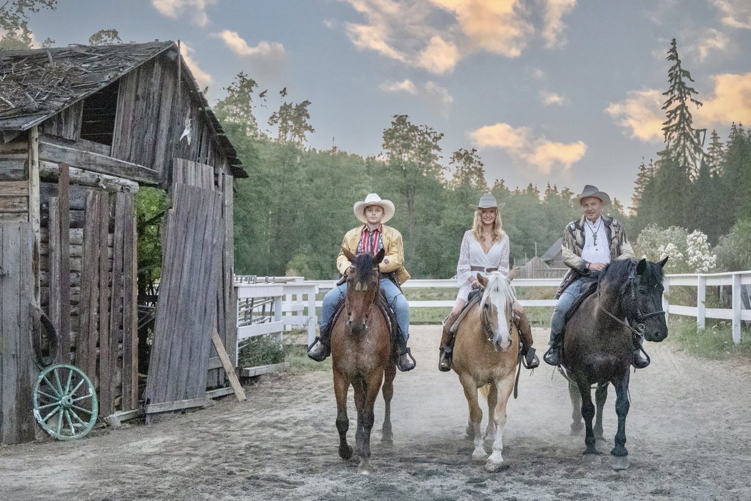 Three family members riding horses beside a barn at sunset