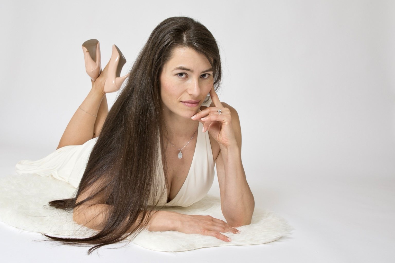 Woman in a white dress lying on her stomach on a soft white rug, resting her hand beside her face